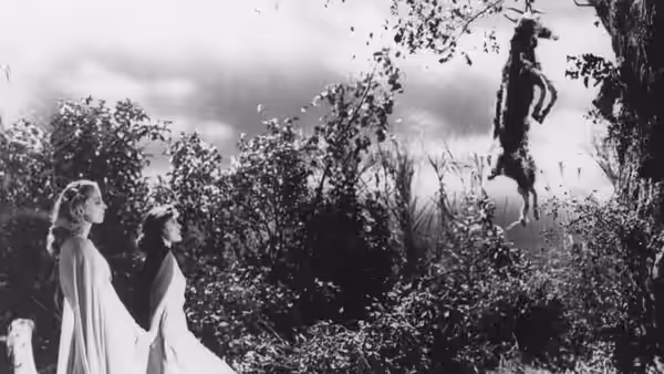 Two women look up at a figure hanging from a tree