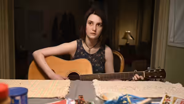 Autumn (Sidney Flanigan) plays an acoustic guitar at a dining table