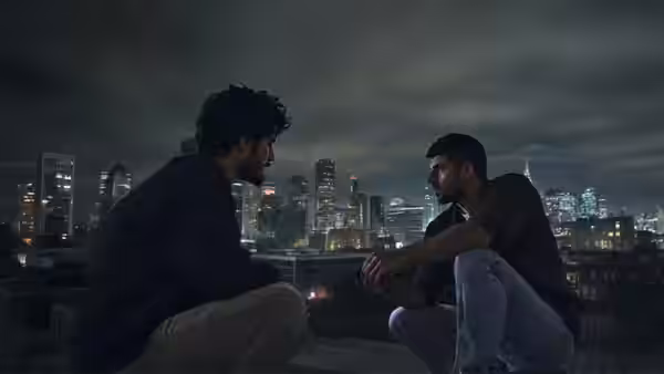 Two men sit facing each other on a rooftop overlooking a city skyline at night