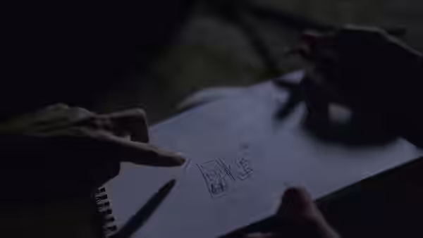 A hand points to a drawing in an open spiral notebook beside a pen