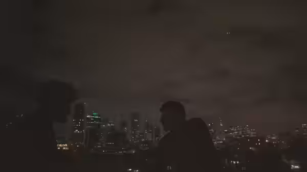 Two men sit on a rooftop with a brightly lit city skyline behind them at night