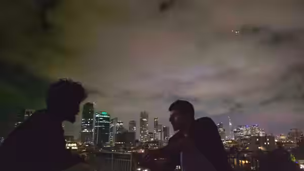 Two men talk beside a rooftop railing overlooking a brightly lit city skyline