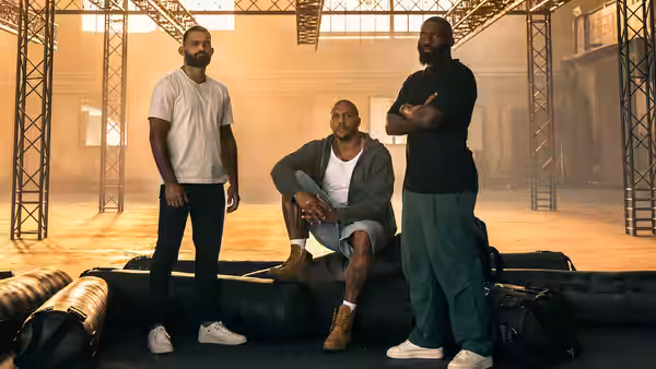Three men stand and sit in a large industrial hall with metal beams