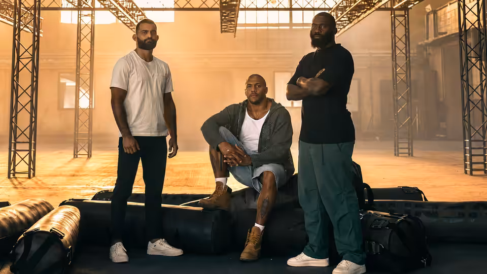 Three men stand and sit in a large industrial hall with metal beams