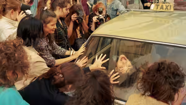 Fans press their hands against a taxi window as a curly-haired man sits inside