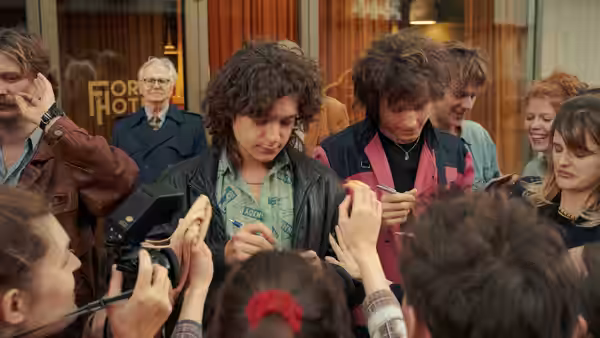 A curly-haired man in a leather jacket signs autographs as fans and photographers crowd around him