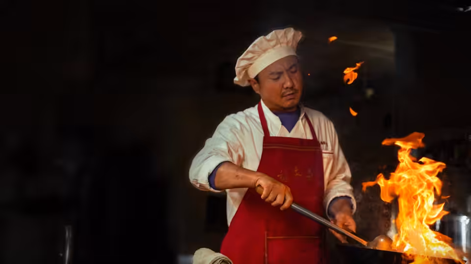 Chef in white hat and red apron stirs a pan over open flames