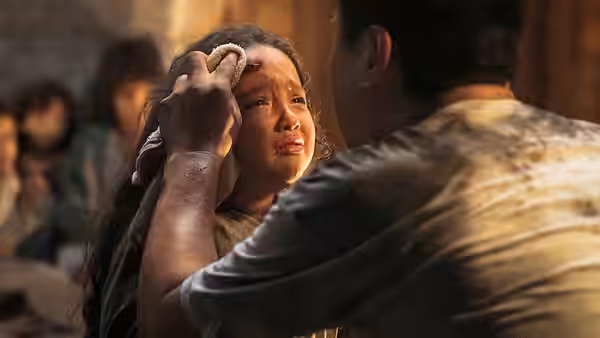 A man presses a cloth to an injured girl's forehead in a crowded shelter