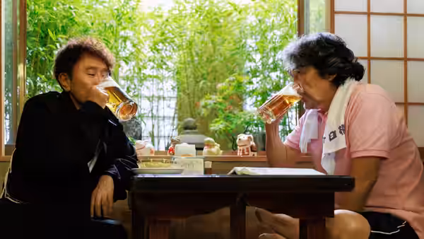 Two men sit across a low table, drinking beer beside a garden window