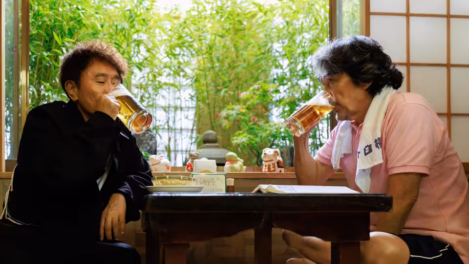 Two men sit across a low table, drinking beer beside a garden window