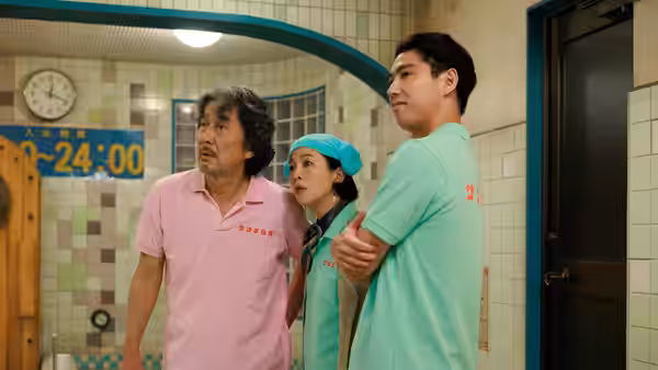 A pink-shirted man, a woman in a scrub cap, and a young man stand in a tiled room