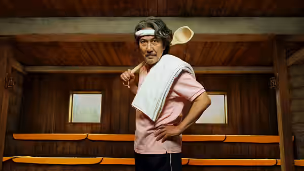 Gen Takasegawa (Koji Yakusho) standing in a wooden bathhouse holding a ladle