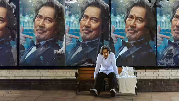 A man sits beneath repeated portraits of himself beside a bench and suitcase