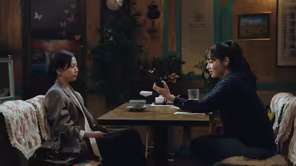 Two women sit across a table as one gestures with her phone