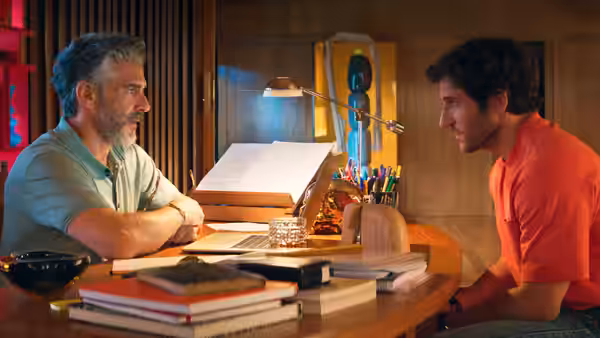 Gray-haired man and younger man in red shirt sit facing each other at desk