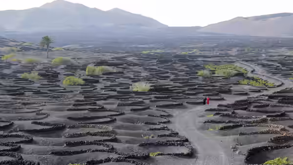 Two figures walk along paths amid vast volcanic stone enclosures and mountains
