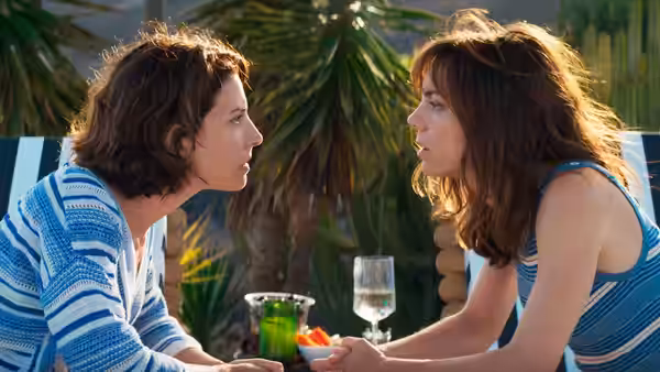 Two women face each other across an outdoor table with drinks and snacks