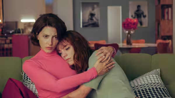 Woman in pink turtleneck embraces another woman on green living room sofa