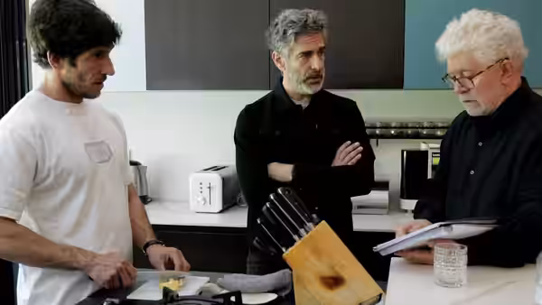 Three men stand around kitchen counter with cutting board and knives