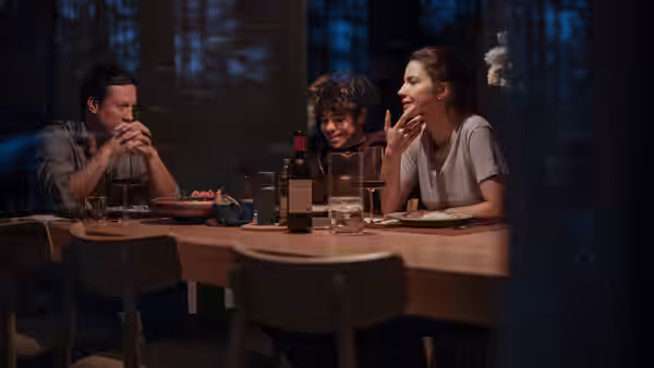 Three people share dinner at a wooden table in a dim forest-side room