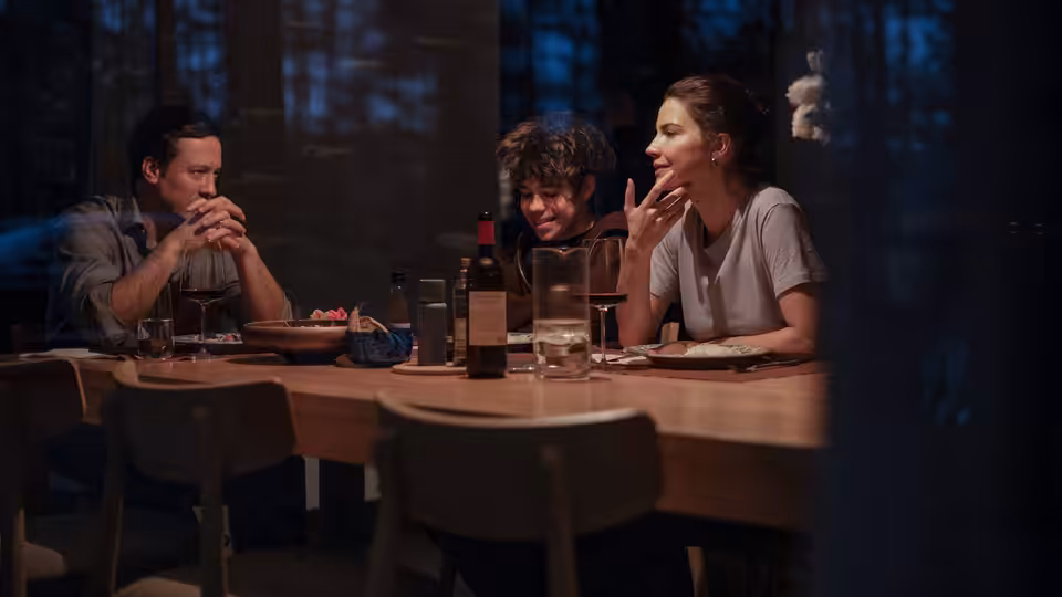 Three people share dinner at a wooden table in a dim forest-side room