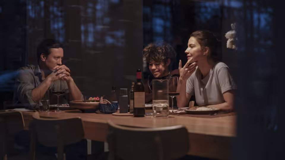 Three people seated at a wooden dining table with wine and food
