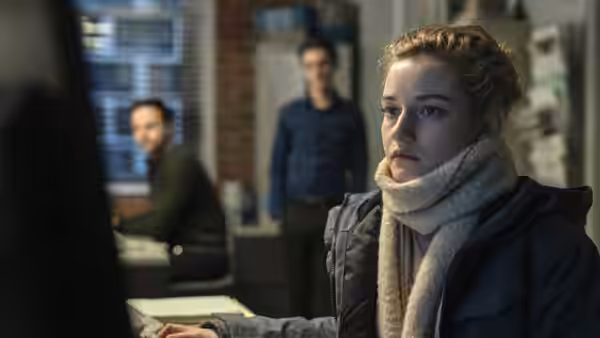 Jane (Julia Garner) sits at her office desk in a coat and scarf, with coworkers behind her