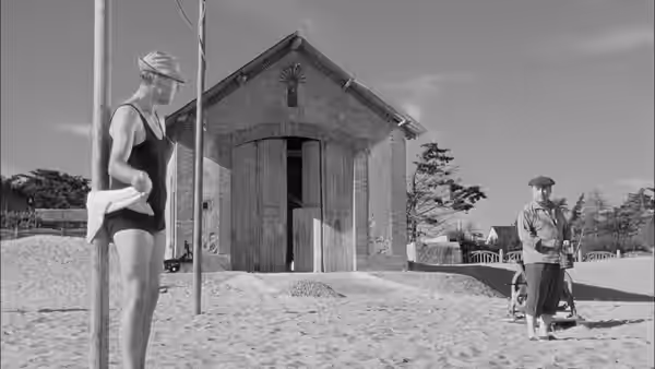 A woman waits beside a beach changing hut as Monsieur Hulot approaches