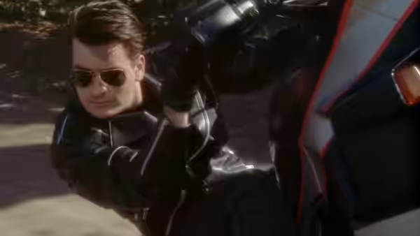 A man in sunglasses and a black leather suit crouches beside a motorcycle