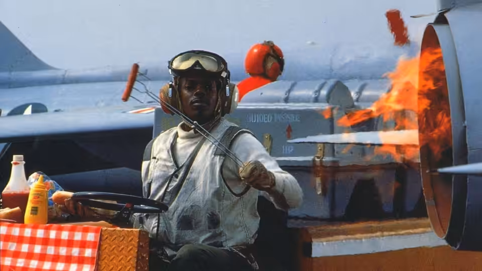 A man in flight gear cooks beside an aircraft as flames engulf its propeller