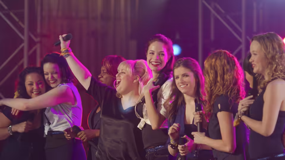 Group of women singing energetically on stage with microphones under colorful lights