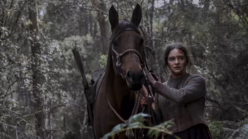 Clare (Aisling Franciosi) stands beside a saddled horse in a forest