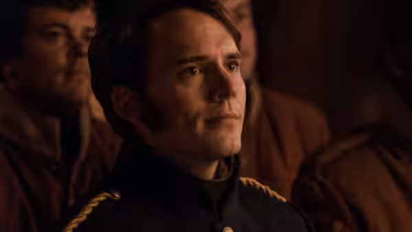 Hawkins (Sam Claflin) stands among soldiers in a dimly lit room