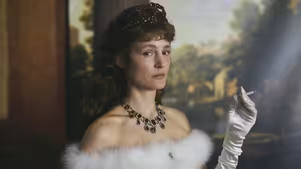 Empress Elisabeth (Vicky Krieps) holds a cigarette in a gloved hand beside a painted backdrop