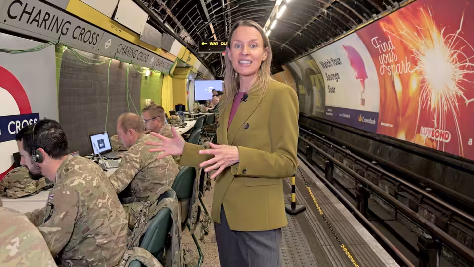 A woman in a green jacket gestures beside uniformed personnel working at control-room computers
