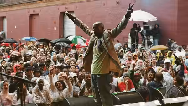 Dave Chappelle with arms raised standing before a massive outdoor crowd