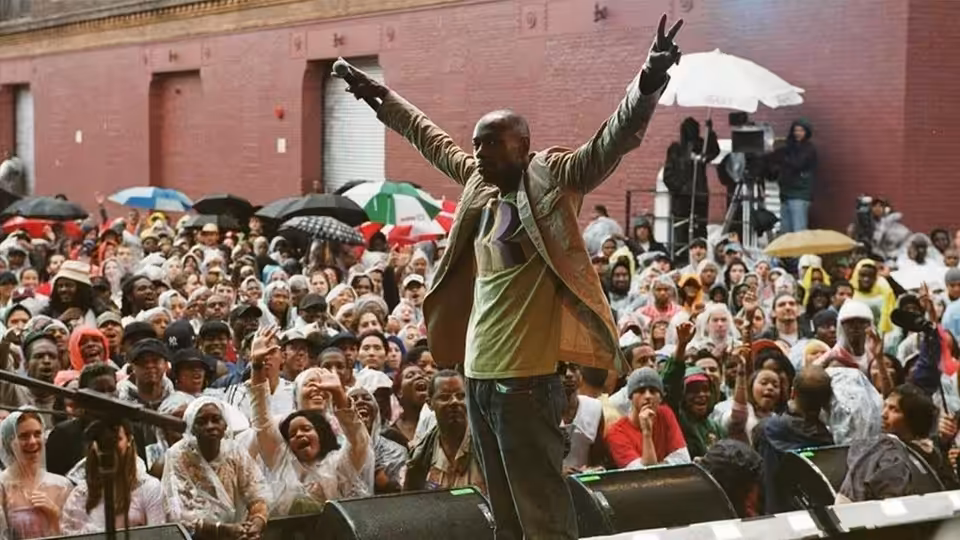 Dave Chappelle with arms raised standing before a massive outdoor crowd