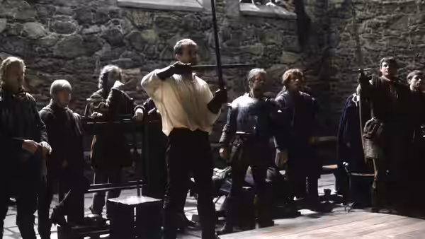 Hamlet (Mel Gibson) holds a sword among courtiers in a stone courtyard