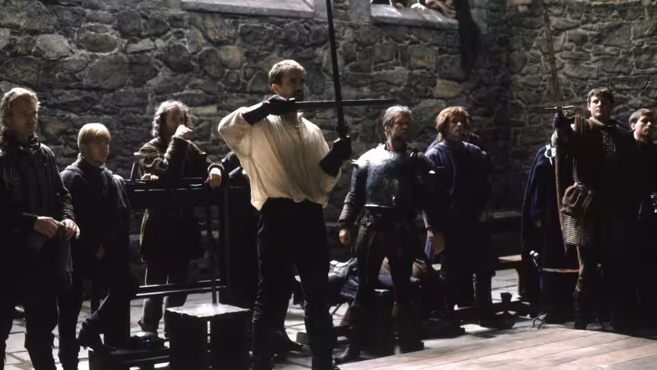 Hamlet (Mel Gibson) holds a sword among courtiers in a stone courtyard