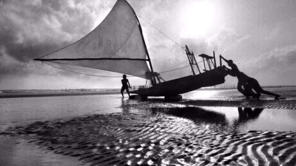 two men with a sailboat on wet sand at the water's edge