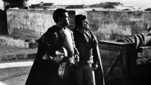 Two men in cloaks and armor stand on a stone balcony by the sea