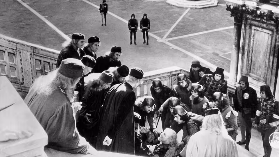 A crowd of men in robes and hats gathered on stone stairs overlooking a courtyard