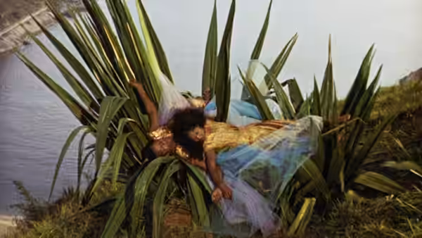 A man supports a woman in a blue dress among broad-leaved plants beside the water