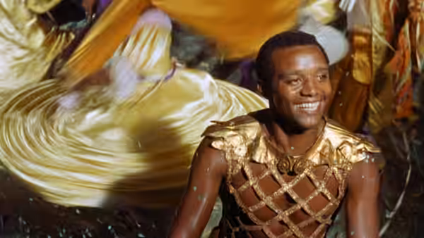 Orfeo (Breno Mello) smiles as he dances in a gold lattice costume