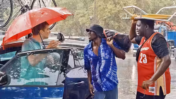 Three men gather around a car during a rainstorm with an umbrella