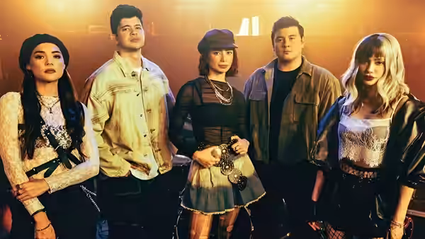 Five young adults in eclectic outfits posing together under warm stage lights