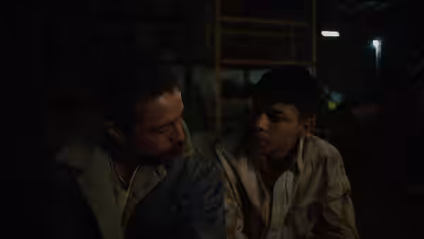 Two men sit side by side in a dimly lit cluttered room at night