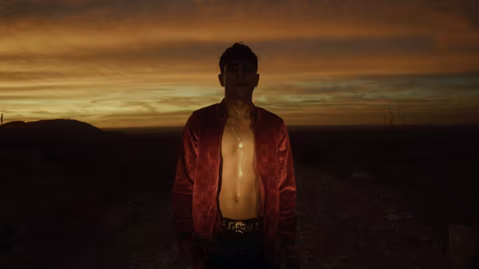 A man in an open red jacket stands shirtless in a desert at dusk