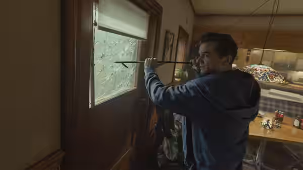 Man in blue hoodie aims crossbow at window of wooden door