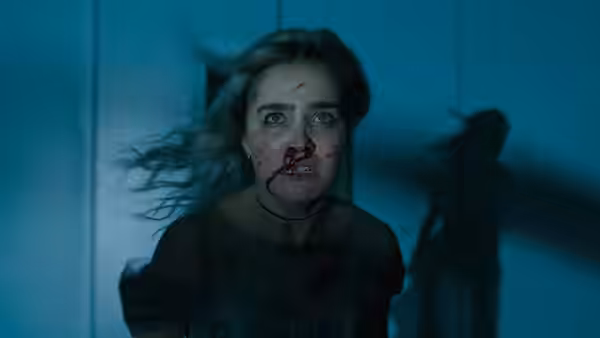Bloodied blonde woman stares forward in a blue-lit hallway